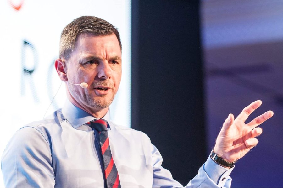 Man speaking with a microphone on stage, wearing a light blue shirt and striped tie, making a gesture.