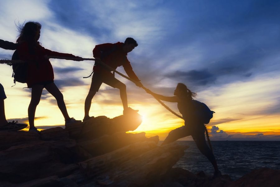 Hikers silhouette helping each other on rocky cliff during sunset, teamwork and adventure concept.