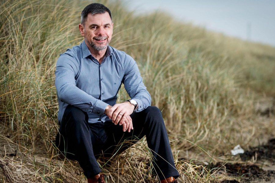 Man sitting on dune grass in relaxed pose, wearing blue shirt and watch, outdoor setting, smiling at camera.