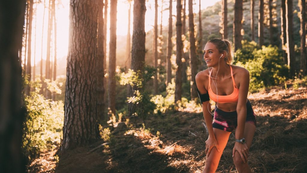 Woman taking a break while jogging in a sunlit forest, wearing sportswear and earphones. Fitness and nature concept.